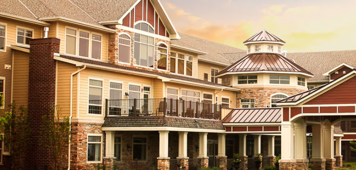 Exterior view of a multi-story senior living facility with beige siding, stone accents, large windows, a balcony with chairs, and a covered entrance with columns under a partly cloudy sky at sunset.