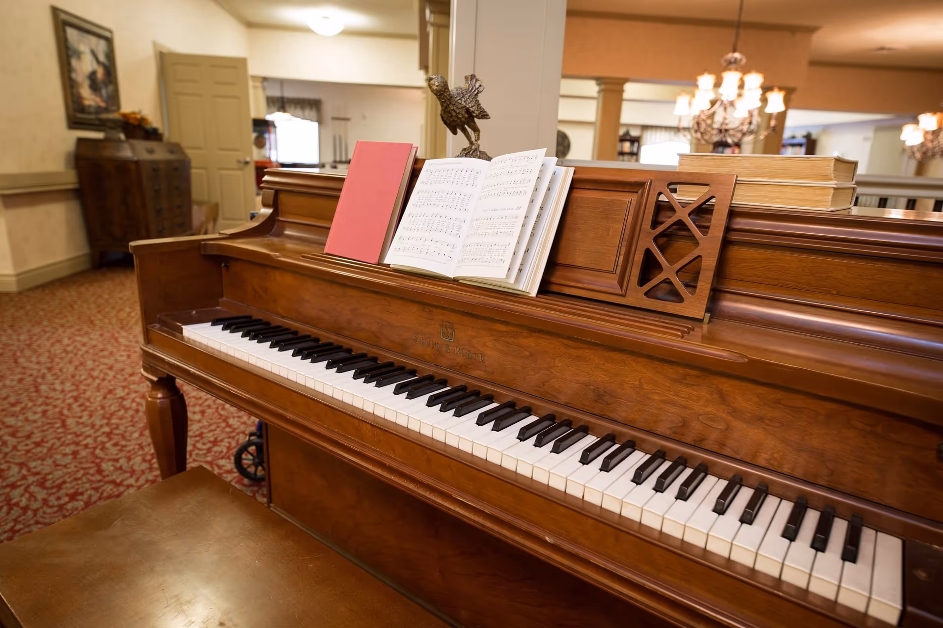 Wooden upright piano with open sheet music and a red book in a carpeted common room with chandeliers and furniture in the background.