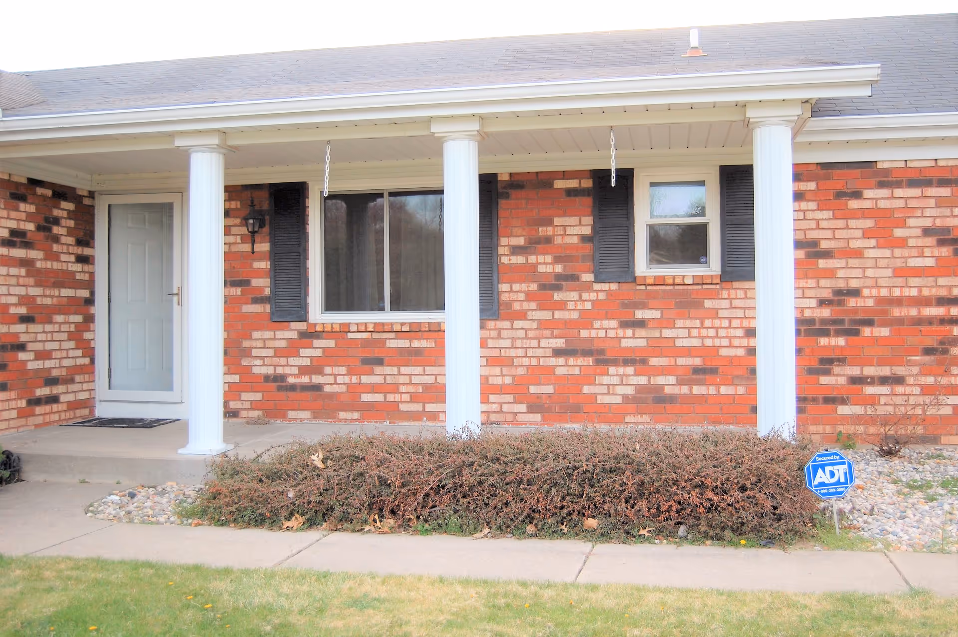 Front exterior view of a brick building with a covered porch supported by white columns. There is a white door on the left, two windows with black shutters, and a small garden bed with shrubs in front. A blue ADT security sign is visible in the garden bed.