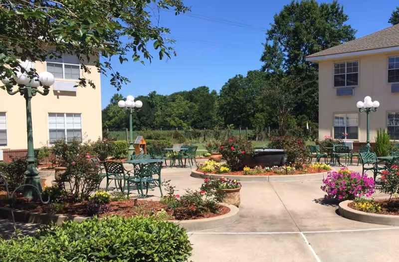 Outdoor patio area at Garden View Assisted Living with green metal tables and chairs, surrounded by flower beds and bushes. Two beige buildings with multiple windows flank the patio, and vintage-style street lamps with globe lights are placed around the area. Trees and a clear blue sky are visible in the background.