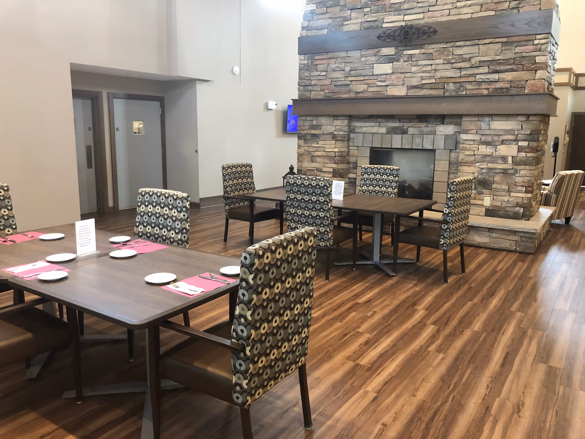 Dining area with set tables and patterned chairs on wood floors in front of a large stone fireplace.