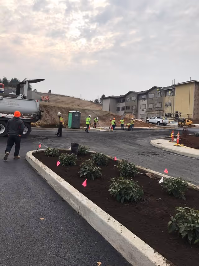 Construction workers in safety vests paving a driveway and planting landscaping in front of a multi-story senior living building under construction.