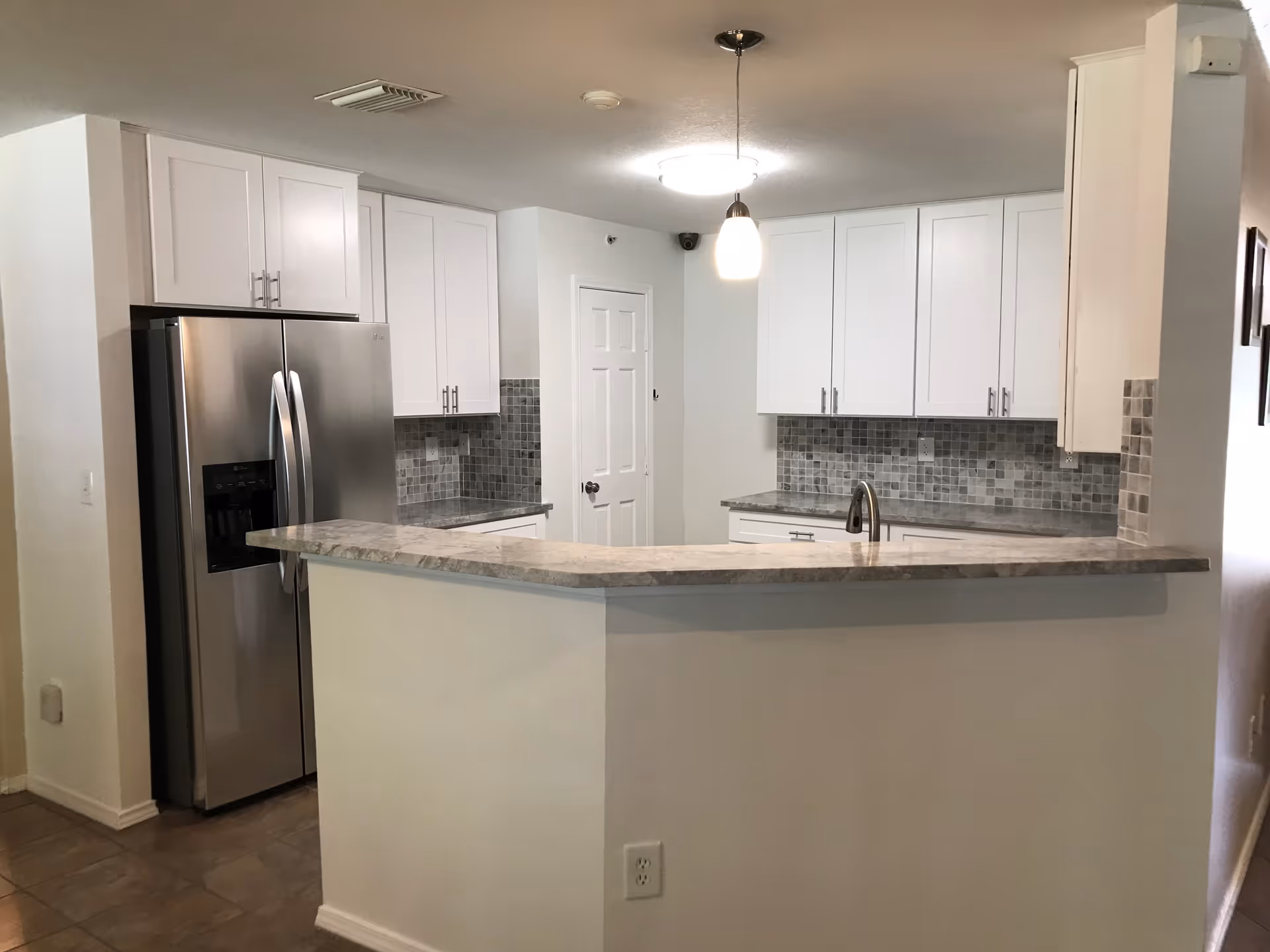 Modern kitchen area with white cabinets, a stainless steel refrigerator, a marble countertop bar, mosaic tile backsplash, and two ceiling lights.