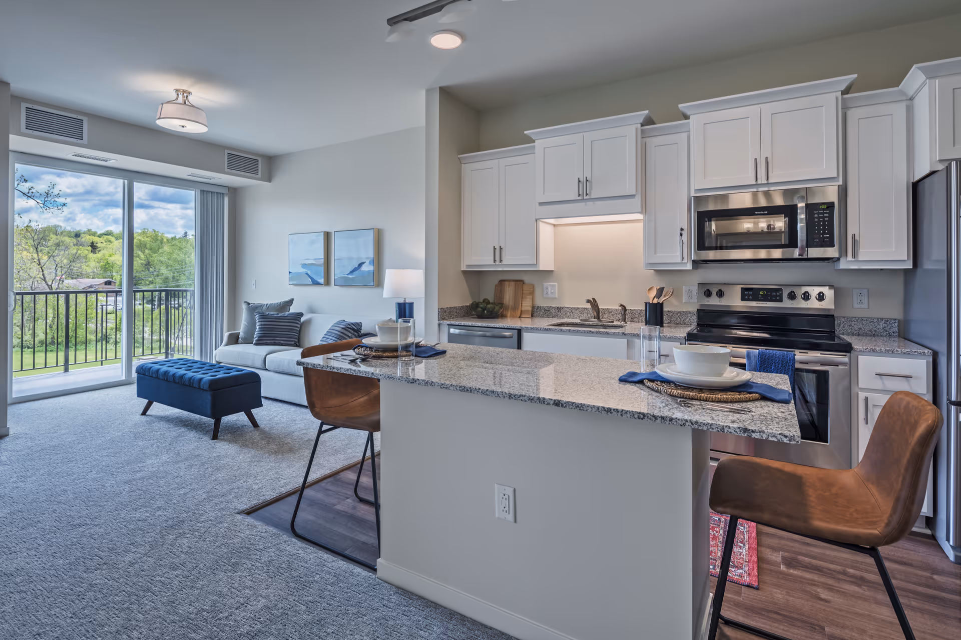 Modern open-concept living space featuring a kitchen with white cabinets, granite countertops, stainless steel appliances, and a breakfast bar with two brown leather chairs. Adjacent to the kitchen is a cozy living area with a white sofa, blue ottoman, and large sliding glass doors leading to a balcony overlooking green trees and a blue sky.