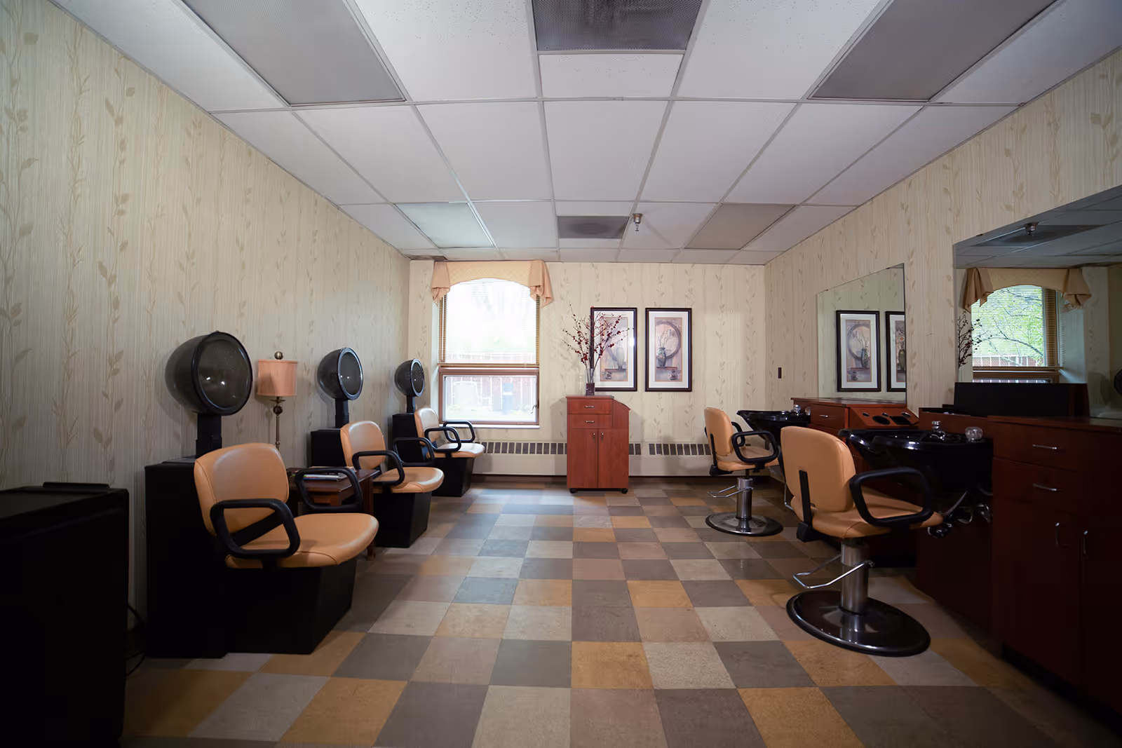 Interior view of a hair salon area in a senior living facility with beige salon chairs, hair drying stations on the left, and hair washing sinks with mirrors on the right. The room has patterned wallpaper, a checkered floor, and two framed pictures on the far wall beneath a window with a valance.