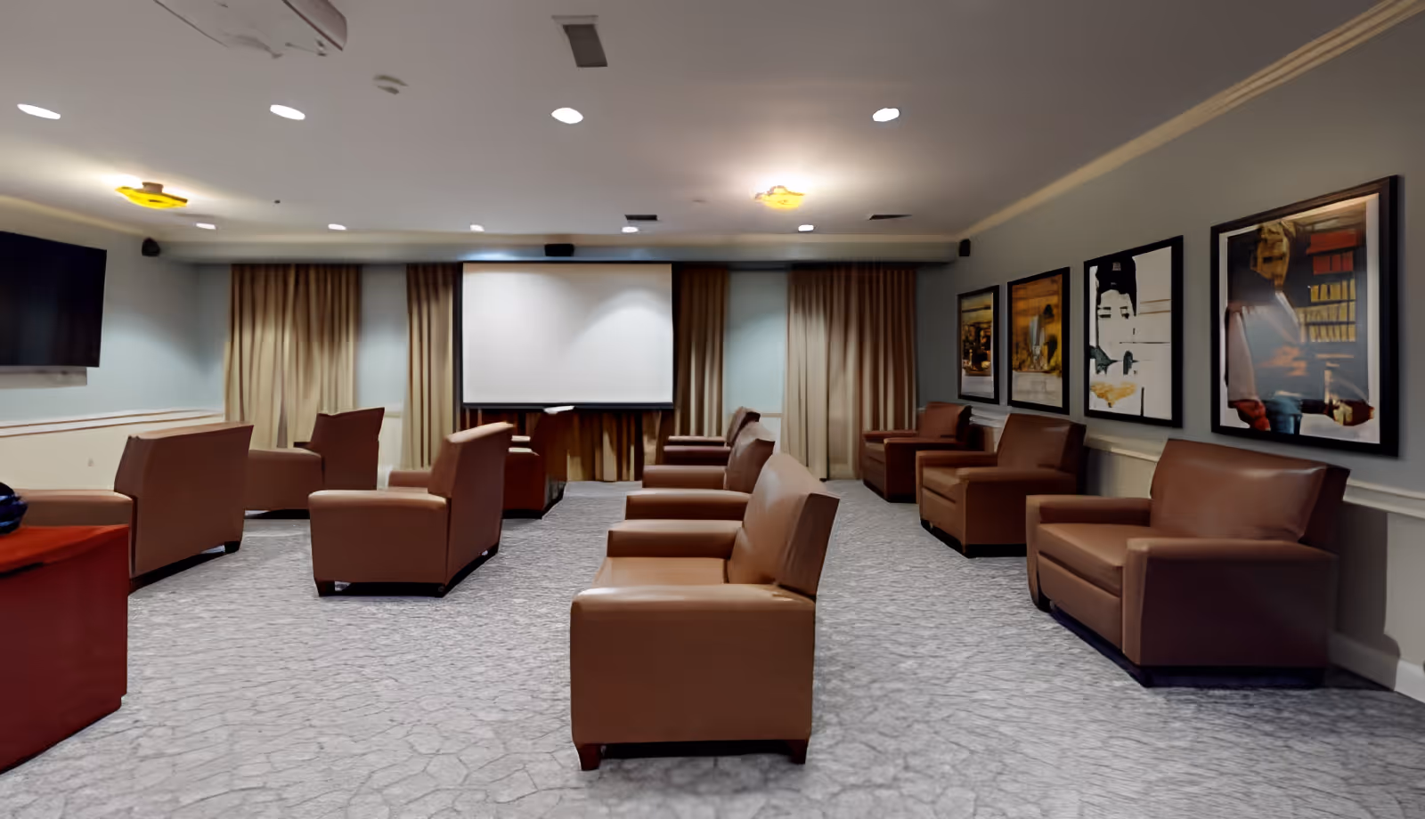 A spacious room with multiple brown leather armchairs arranged in rows facing a large projection screen. The room has light-colored walls with framed artwork on one side and beige curtains covering windows at the far end. The floor is carpeted in a light gray pattern, and the ceiling has recessed lighting and two ceiling-mounted light fixtures.
