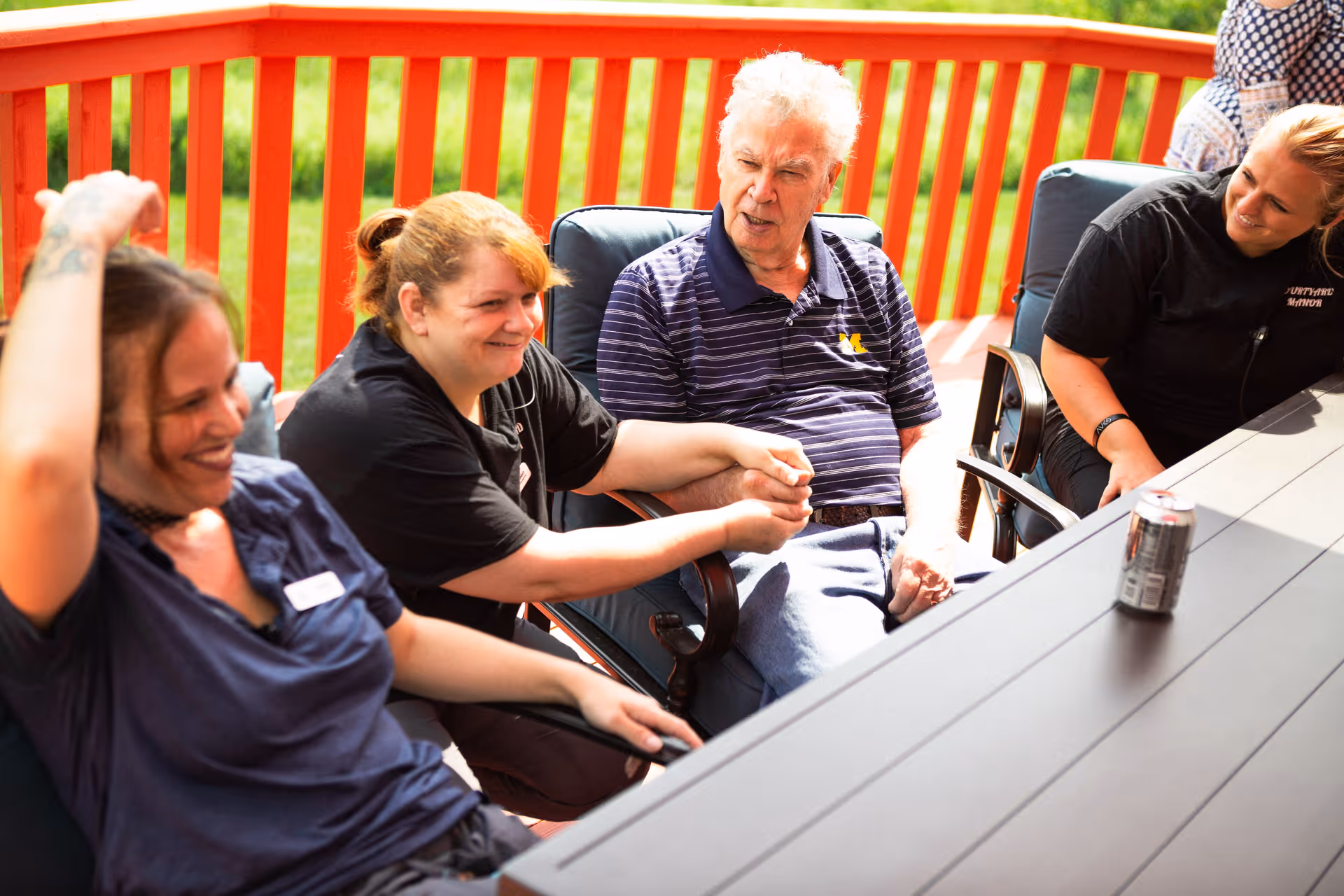 A group of three women and one elderly man sitting around a table on an outdoor deck with a red railing, engaging in conversation and smiling. There is a can of soda on the table.
