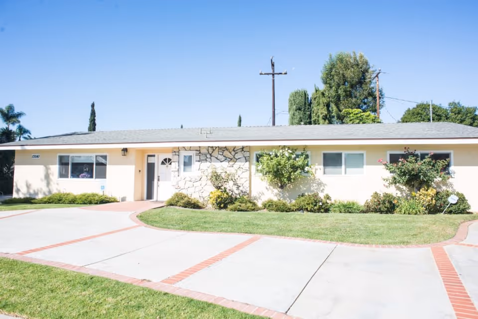 Single-story ranch-style building front with a driveway, lawn and shrubs under a clear blue sky.