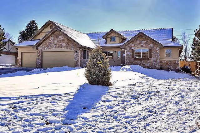 A single-story stone and brick house with a three-car garage, surrounded by snow-covered ground and a small evergreen tree in the front yard under a clear blue sky.