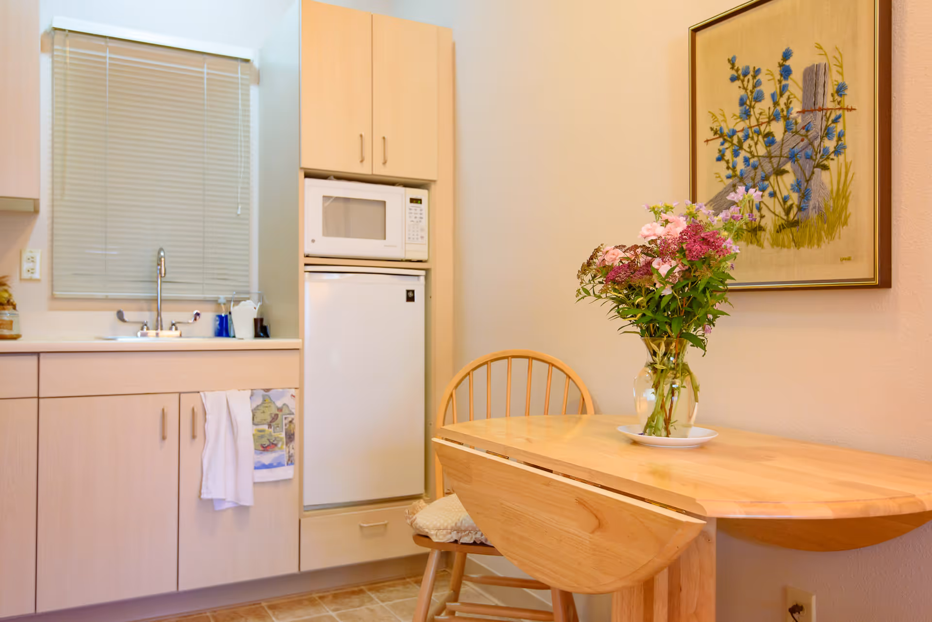 Small kitchenette with a sink, microwave, mini-fridge and a wooden table topped with a vase of flowers.
