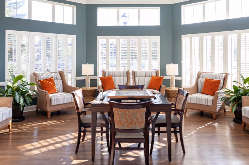 Bright and airy common area with large windows featuring white shutters, several cushioned armchairs with orange throw pillows, a wooden table with chairs arranged around it, and green potted plants on either side.