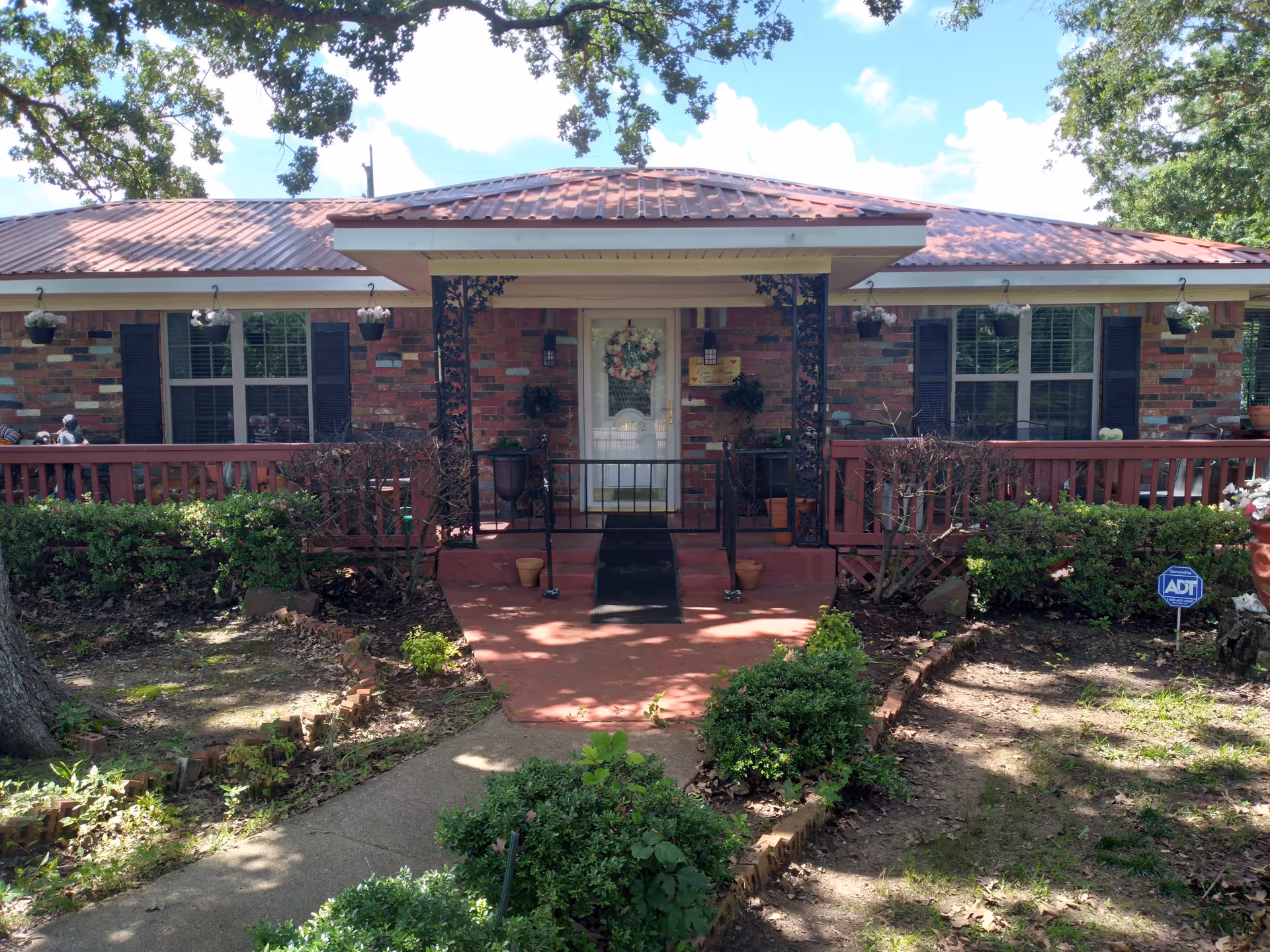 Front exterior view of a single-story brick building with a metal roof and a covered porch entrance. The porch has decorative black metal supports and a white door with a floral wreath. There are windows with black shutters on either side of the door, hanging flower pots, and trimmed bushes along the walkway leading to the entrance. A blue ADT security sign is visible in the garden area to the right.