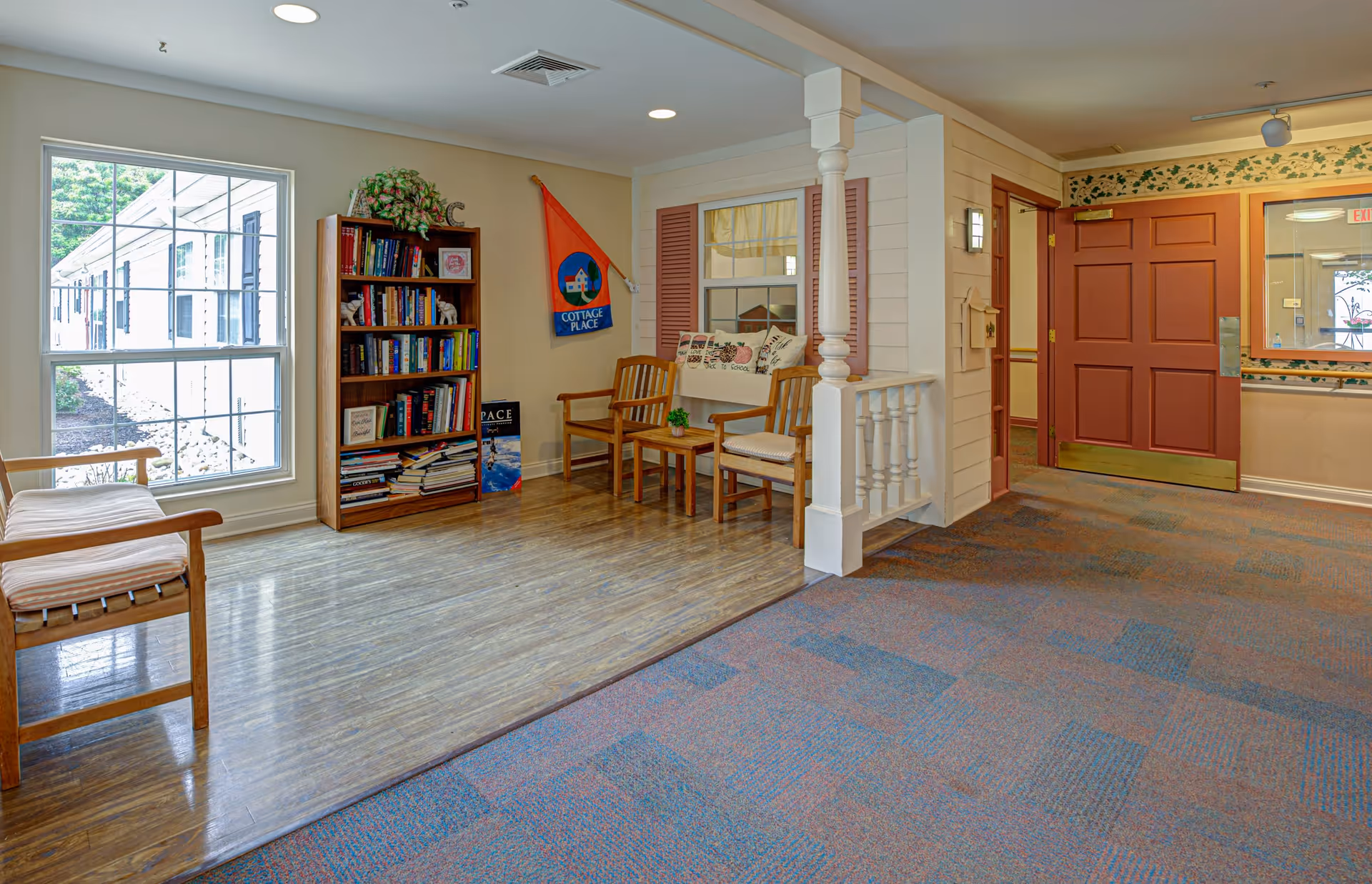 A cozy interior common area with wooden chairs, a small table, a bookshelf filled with books, and a large window letting in natural light. The area has a mix of wood and carpet flooring, and a red door is visible in the background.