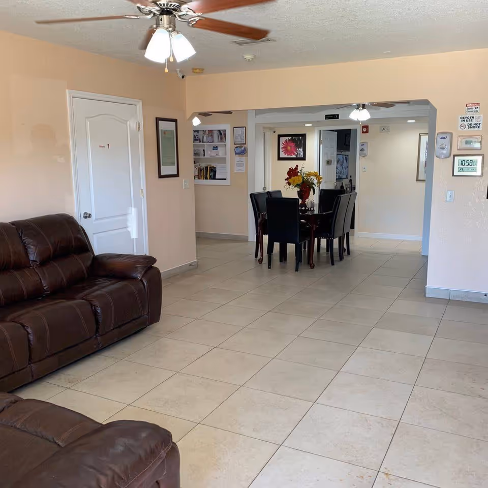 Interior view of a senior living facility showing a tiled floor with brown leather sofas on the left and a dining area with a table and six chairs in the background. The walls are light peach, and there is a ceiling fan with lights. Various wall decorations and signs are visible, including a door labeled 'Room 1'.
