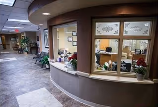 Interior view of a senior living facility hallway with a curved reception desk window. The hallway has tiled flooring, potted plants, and seating areas along the walls. The reception area behind the window has office furniture and decorations.