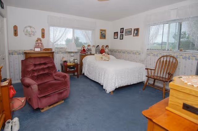 A cozy bedroom with a single bed covered in a white quilt, several decorative dolls placed on the headboard, a burgundy recliner chair, a wooden rocking chair, and a small wooden nightstand with a lamp. The room has blue carpet, floral wallpaper border, and two windows with white lace curtains letting in natural light. Various framed pictures and decorations adorn the walls.