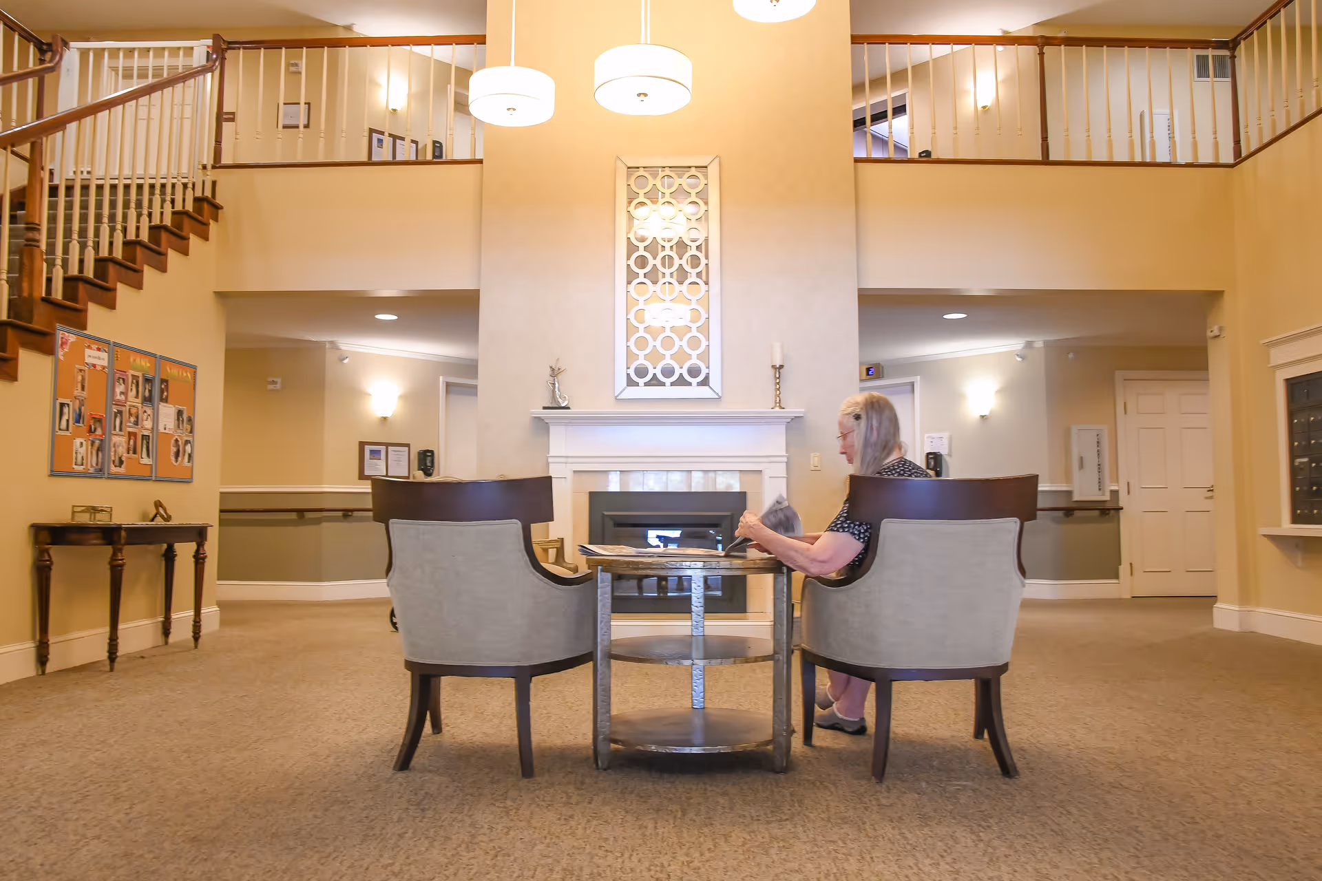 A senior woman sitting at a small round table with two chairs in a spacious, well-lit common area of a senior living facility. The room features a fireplace with a decorative mirror above it, two hanging pendant lights, a staircase with wooden handrails, and bulletin boards on the wall.
