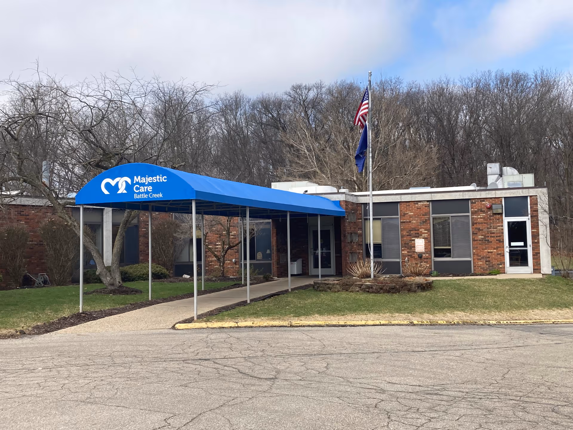 Exterior view of Majestic Care of Battle Creek building with a blue canopy entrance, American and state flags on a flagpole, leafless trees, and a paved driveway in front.