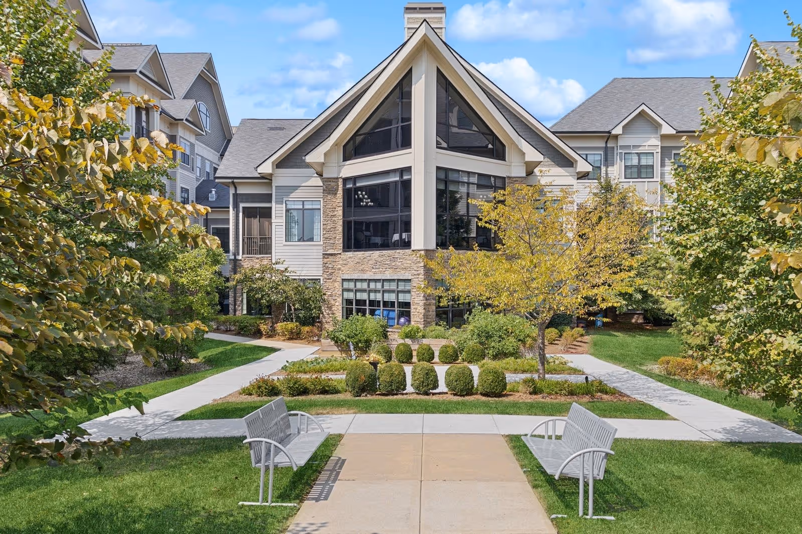 Exterior view of a senior living facility building with large windows and a peaked roof, surrounded by well-maintained landscaping including trees, bushes, and green grass. Two metal benches face each other on a concrete pathway leading to the building entrance.