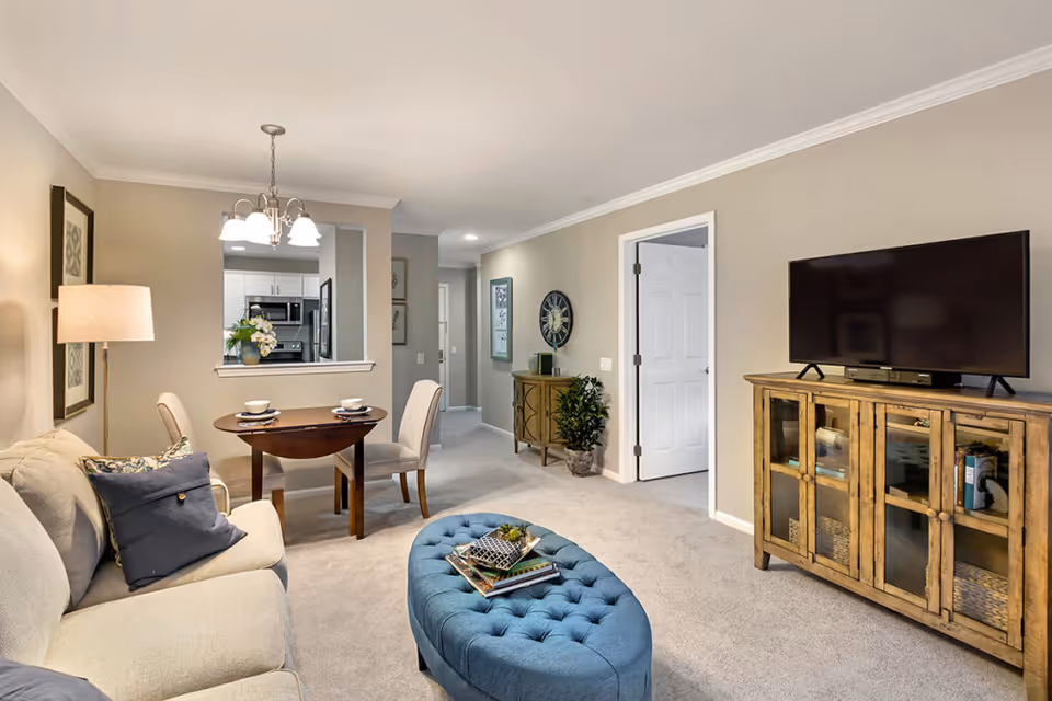 A cozy living room and dining area in a senior living facility with beige walls and carpet. The living room features a beige sofa with cushions, a blue tufted ottoman with books and decor on top, and a wooden cabinet with a flat-screen TV. The dining area has a small round wooden table set for two with chairs, and a hanging light fixture above. The kitchen is visible through a pass-through window, and a hallway leads to other rooms.