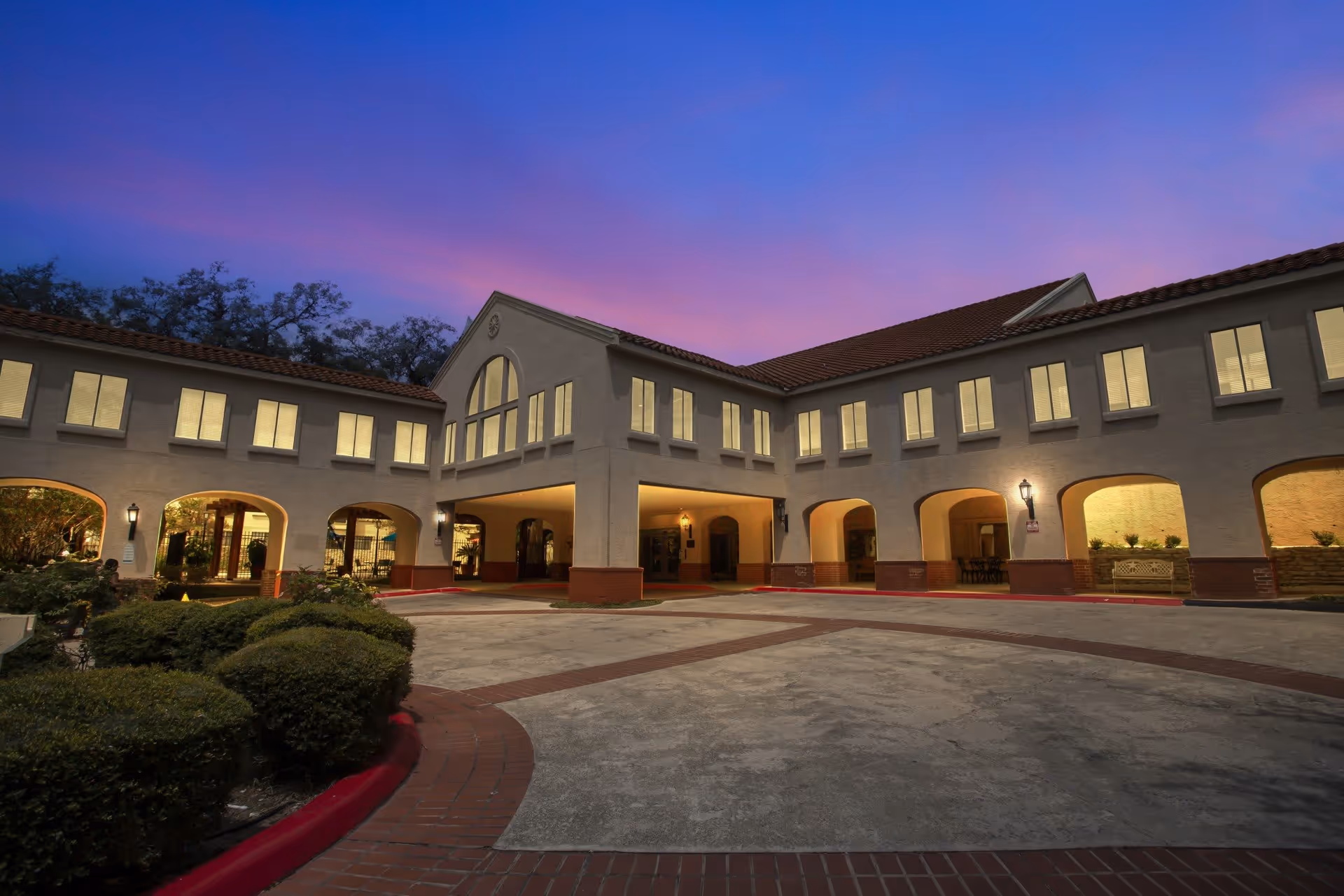 Exterior view of Madison Estates senior living facility at dusk, showing a two-story building with illuminated windows, arched entryways, and a circular driveway surrounded by landscaped bushes.