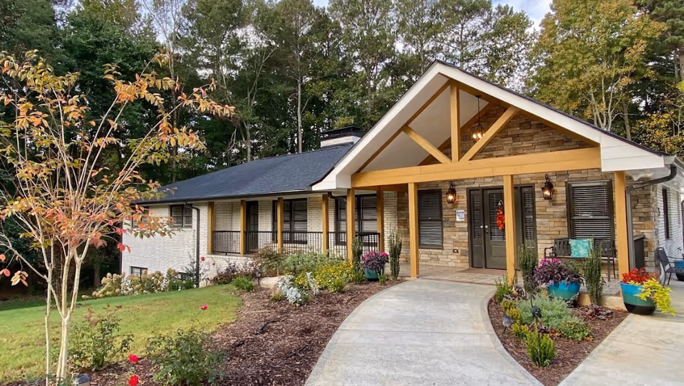 Front exterior view of a senior living facility named Golden Manor Senior Living, featuring a covered entrance with wooden beams, stone and brick facade, a curved concrete walkway, and landscaped garden beds with various plants and flowers. Trees and greenery surround the building.