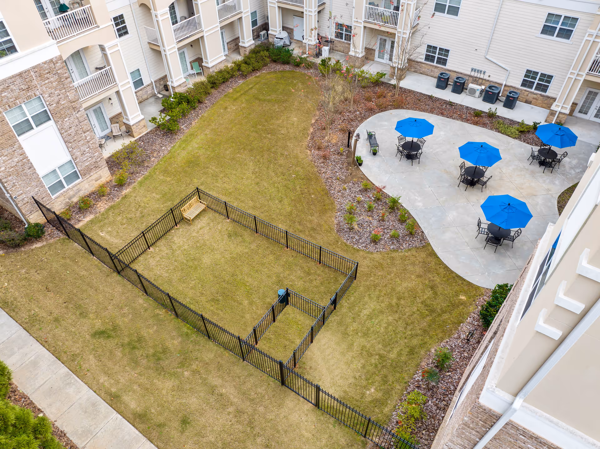 Aerial view of an outdoor courtyard area in a residential facility with a fenced grassy section containing a wooden bench, a concrete patio with several black metal tables and chairs shaded by blue umbrellas, surrounded by landscaped plants and the building's exterior walls with windows and balconies.