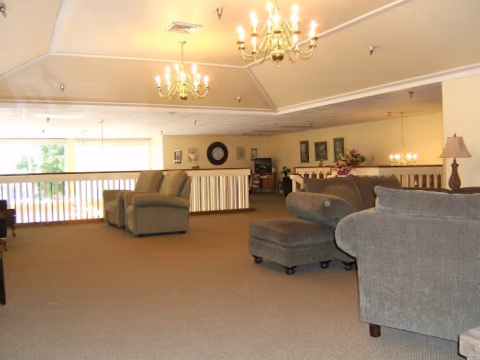 A spacious living room area in a senior living facility with beige carpet, multiple gray upholstered armchairs and sofas, a matching ottoman, and decorative chandeliers hanging from the ceiling. The room has a railing overlooking a lower level and framed artwork on the walls.