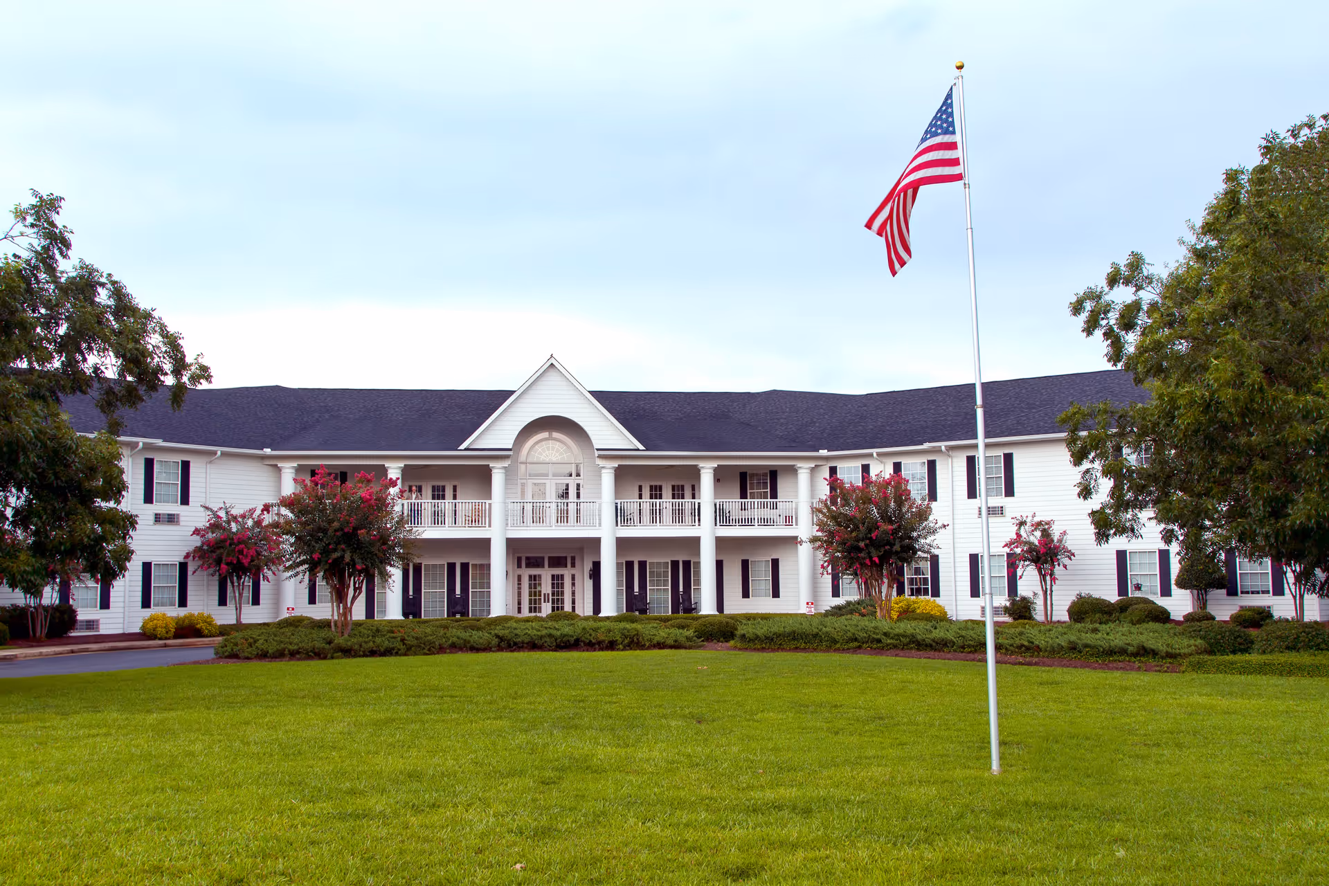 Front exterior view of a large two-story white building with black shutters, a central balcony, and an American flag on a flagpole in the foreground, surrounded by green grass and trees.