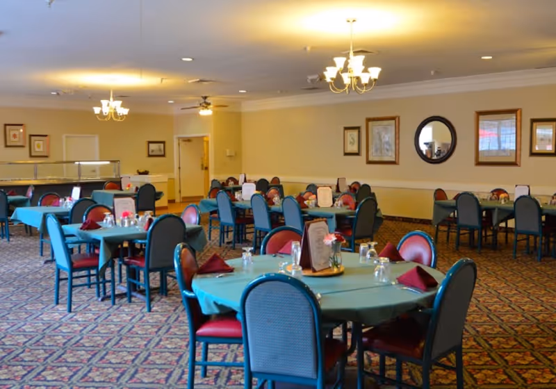 A spacious dining room with multiple tables covered in teal tablecloths, each set with glasses, napkins, and menus. The room has patterned carpet flooring, beige walls adorned with framed pictures and a round mirror, and ceiling lights including chandeliers and ceiling fans.