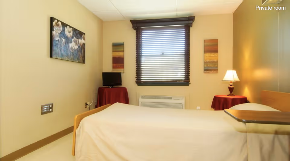A private senior facility bedroom with a single bed, window with blinds, small TV, and a bedside lamp on a round table.