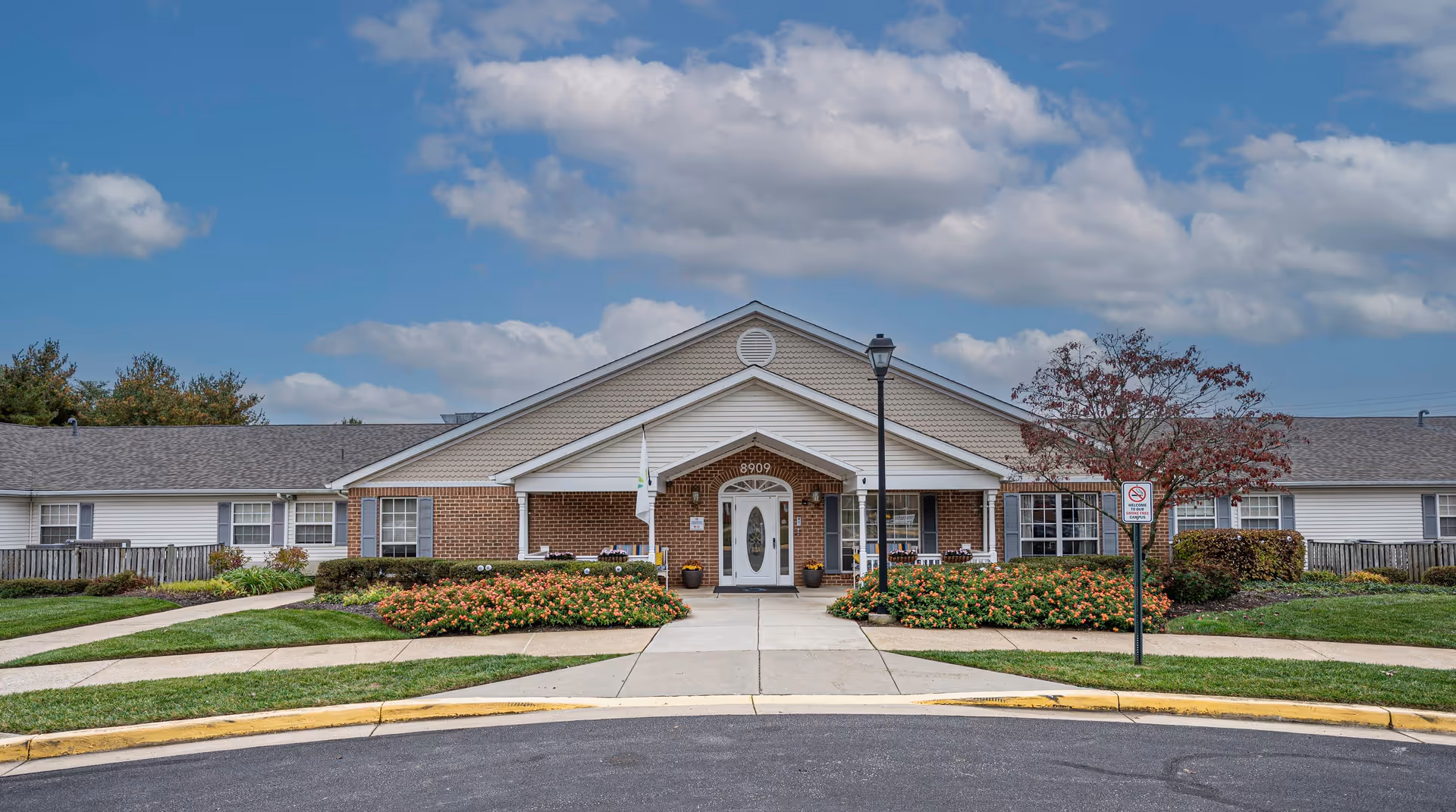 Front exterior view of Arden Courts - ProMedica Memory Care Community in Pikesville, showing a single-story brick and siding building with a peaked roof, white entrance door, landscaped flower beds, a sidewalk, and a lamppost under a partly cloudy sky.