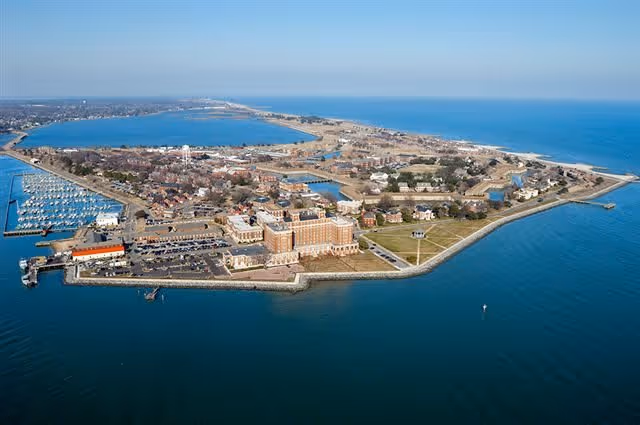 Aerial view of a coastal area featuring a large building complex surrounded by water on three sides, with a marina on the left and open sea on the right. The area includes parking lots, green spaces, and various smaller buildings.