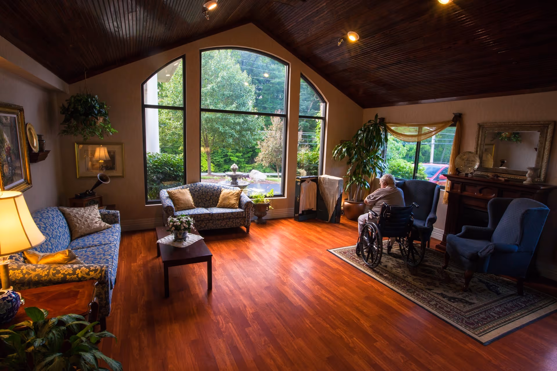 A cozy living room with wooden floors and a high wooden ceiling. The room features two blue patterned sofas, a small wooden coffee table with a flower arrangement, a large window with a view of greenery and a fountain outside, and a fireplace with a mirror above it. An elderly person in a wheelchair is seated near the window, facing outside. There are also two blue armchairs on a rug near the fireplace and several plants around the room.