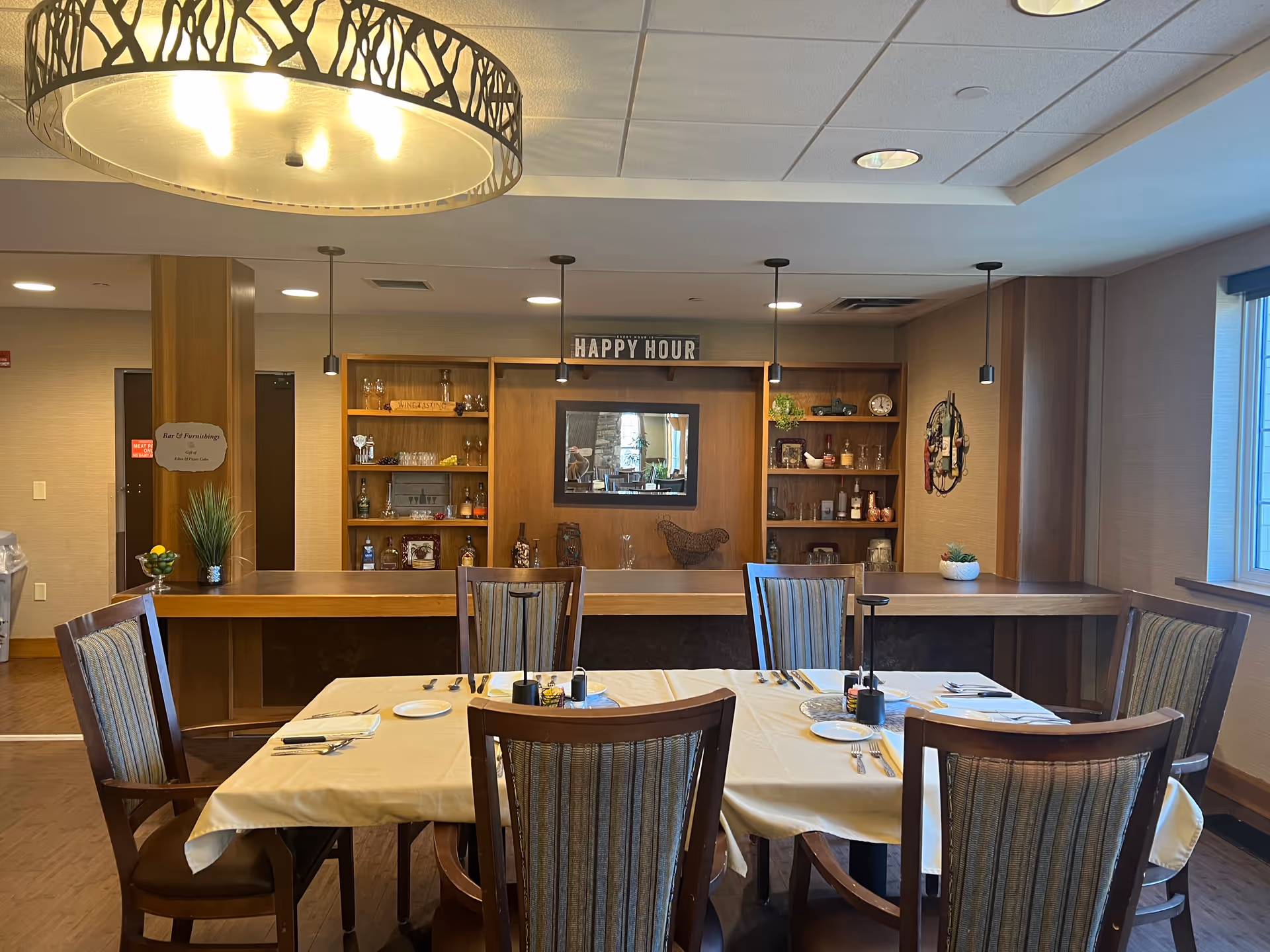 Dining room with a set table and wooden chairs facing a bar area with shelving and a 'Happy Hour' sign.
