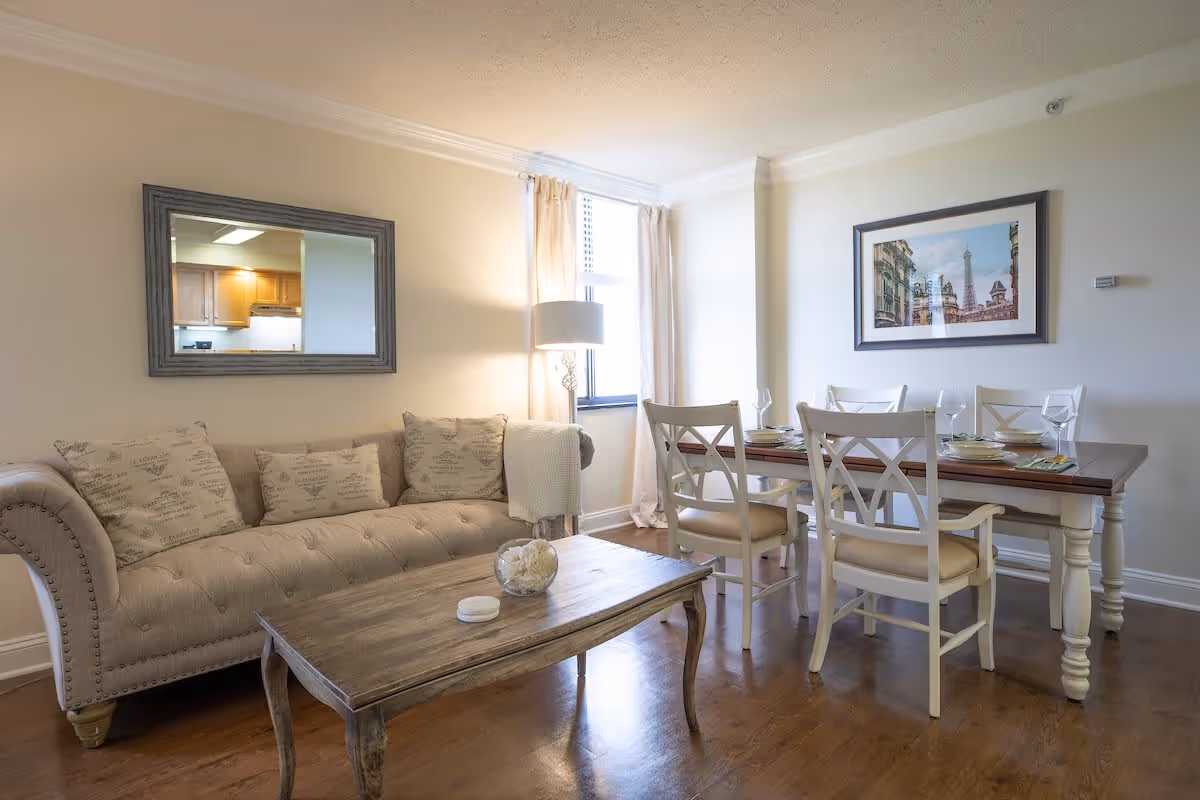 A cozy living and dining area featuring a beige tufted sofa with patterned cushions, a wooden coffee table with decorative items, a dining table set with four white chairs and place settings, a floor lamp near a window with light curtains, a large mirror reflecting a kitchen, and a framed picture on the wall.
