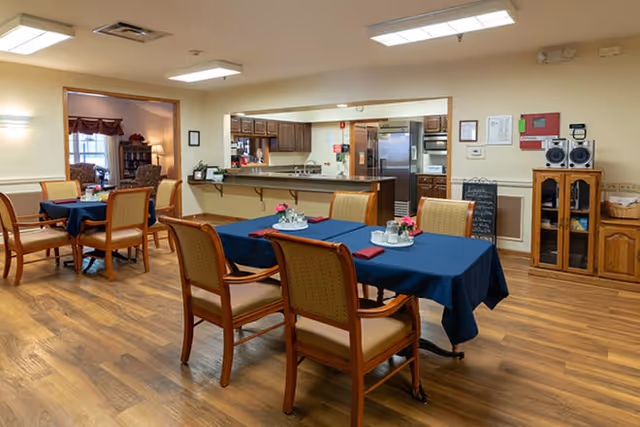 A dining area in a senior living facility with wooden floors and tables covered with blue tablecloths. Each table has chairs with cushioned seats and backs, and small flower arrangements with napkins. In the background, there is an open kitchen with wooden cabinets, a refrigerator, and a counter. The room is well-lit with ceiling lights and has a cozy, welcoming atmosphere.
