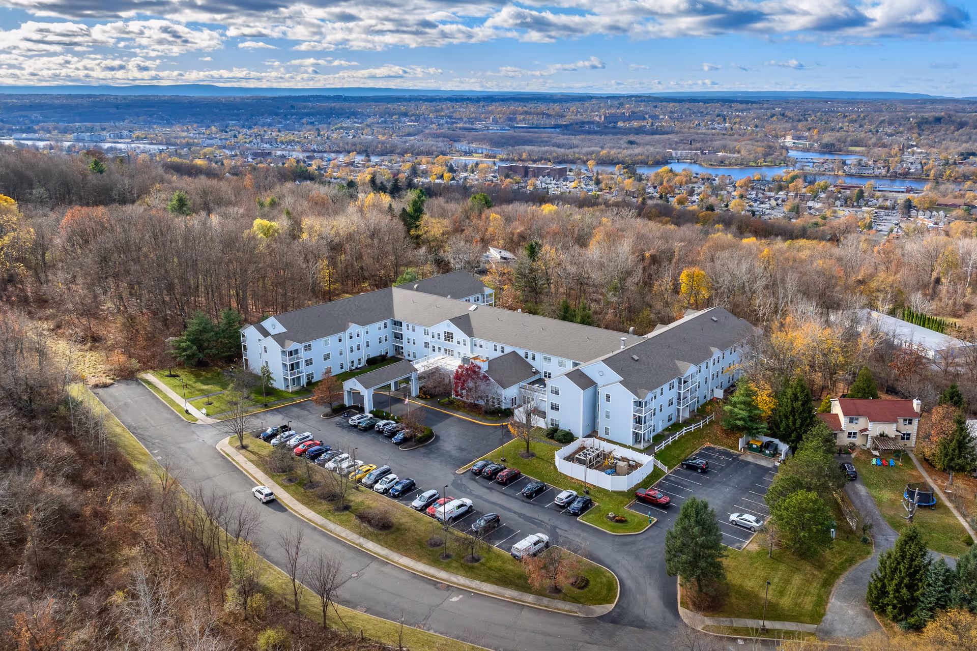 Aerial view of Diamond Ridge, a large senior living community building surrounded by trees with autumn foliage. The building is white with a gray roof and has multiple parking lots with cars parked. The surrounding area includes wooded land and a distant view of a town with water bodies and hills under a partly cloudy sky.