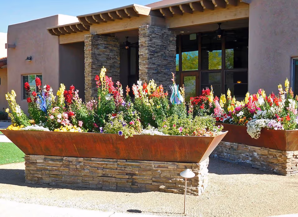 Stone-faced building entrance with large rust-colored raised planters overflowing with colorful flowers.