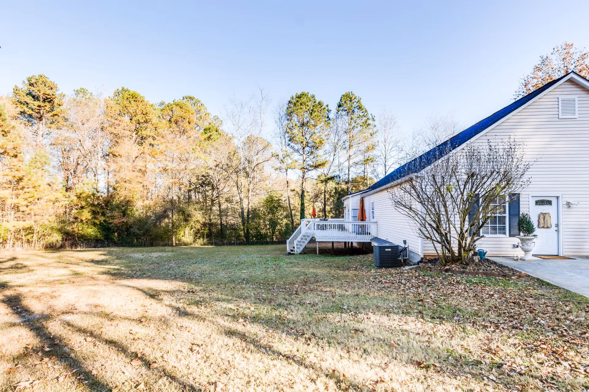 A white residential building with a small deck and stairs leading to a grassy yard surrounded by trees under a clear blue sky.