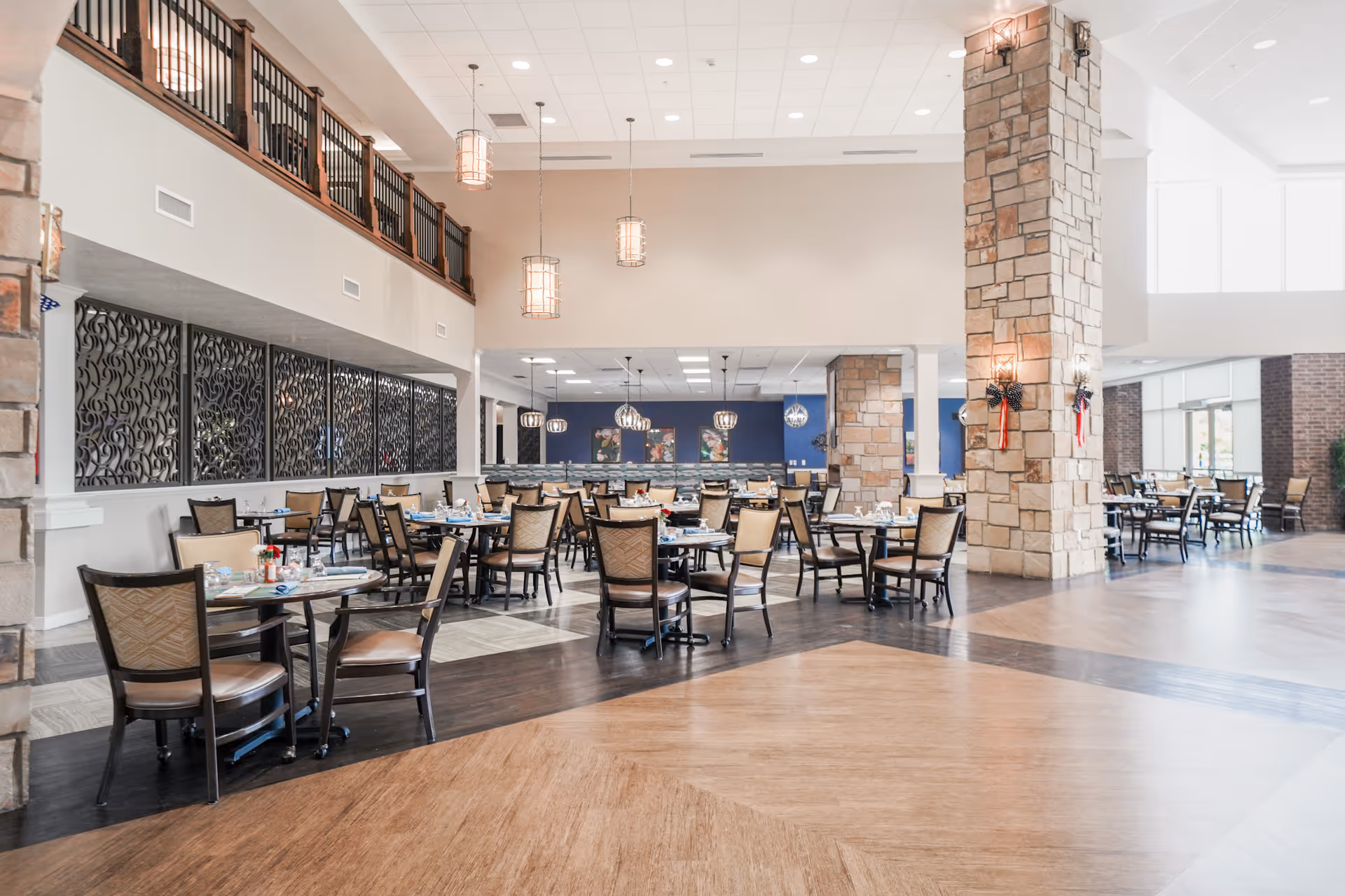 Spacious dining room in a senior living facility with multiple tables and chairs arranged neatly. The room features high ceilings with hanging pendant lights, stone pillars, decorative wall panels, and large windows allowing natural light to fill the space.