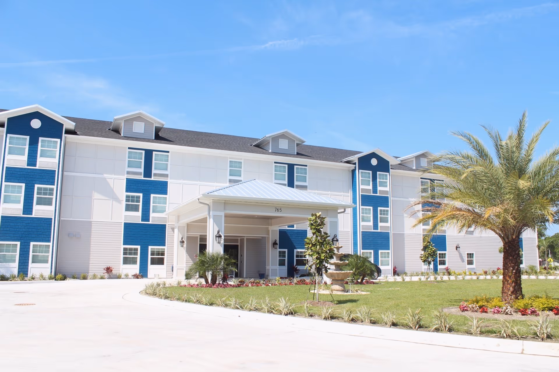 Front exterior view of a three-story senior living facility building with blue and white siding, a covered entrance, landscaped lawn with palm trees and shrubs, under a clear blue sky.