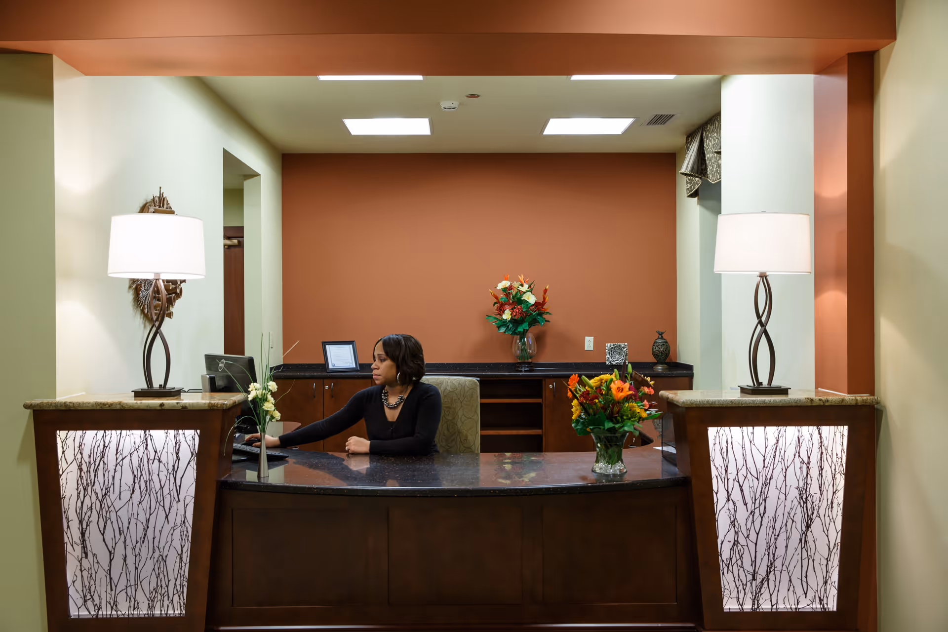 Reception area with a woman sitting behind a dark wood curved desk, working on a computer. The desk has two decorative lamps with white shades on either side and floral arrangements on top. The background wall is painted a warm brown color with additional flowers and decor on a counter behind the desk.