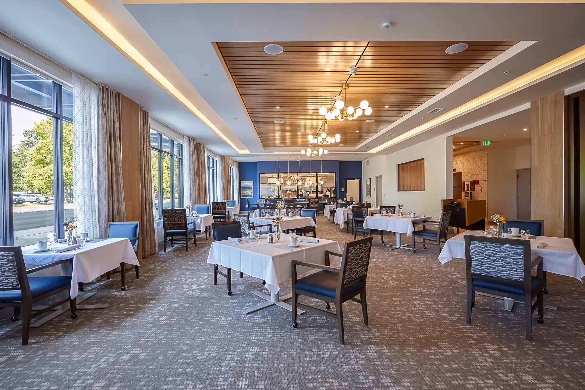 A spacious dining room with multiple tables covered in white tablecloths, each set with cups, glasses, and small flower arrangements. The room features large windows with curtains on the left side, allowing natural light to fill the space. The ceiling has a wooden panel design with modern light fixtures hanging down. The back wall is painted blue and has a serving area with pendant lights and staff visible behind the counter.