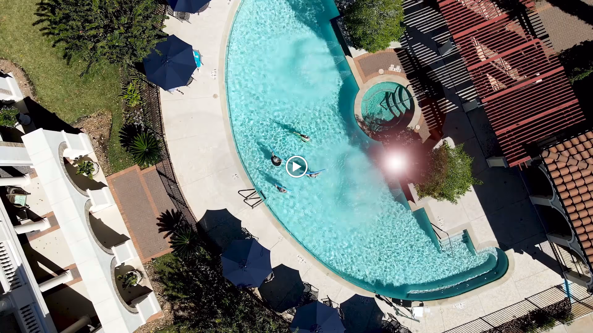 Aerial view of an outdoor swimming pool area with several people swimming and relaxing in the water. The pool is surrounded by a concrete deck with multiple dark blue umbrellas providing shade over lounge chairs. There is a hot tub adjacent to the pool and a shaded pergola structure nearby. Green grass and landscaping are visible around the pool area.