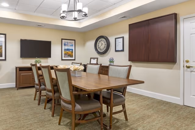 Well-lit communal dining room with a rectangular wooden table surrounded by chairs, a wall-mounted TV, and a large clock.