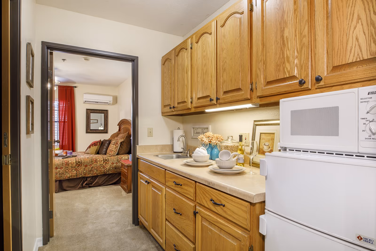 View of a small kitchen area with wooden cabinets, a countertop with dishes and decorative items, a microwave on top of a mini refrigerator, and a sink. Through an open doorway, a bedroom with a bed, pillows, a nightstand, a mirror, and red curtains is visible.