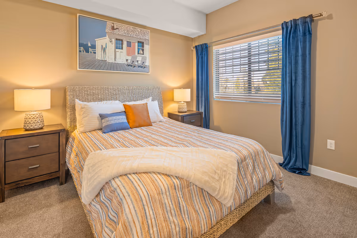 Bright, cozy bedroom with a striped bed, woven headboard, two bedside tables with lamps, a window with blue curtains, and beach-themed wall art.