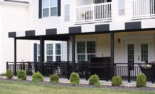 Ground-floor patios and upper balconies of a light-colored senior living building with black railings and small shrubs.