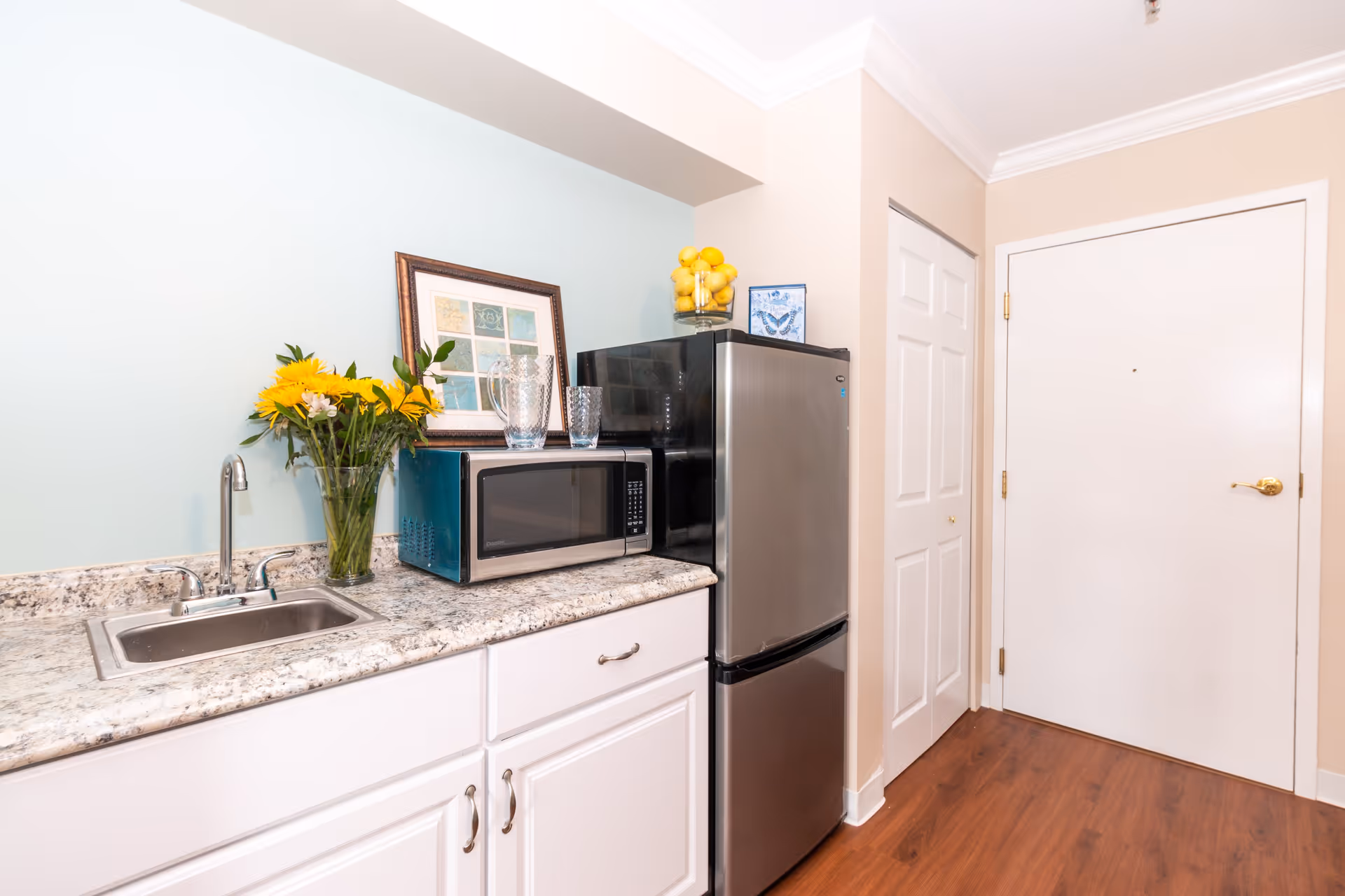 A small kitchen area with a stainless steel sink, a microwave, and a stainless steel refrigerator. There is a vase with yellow flowers on the countertop, a framed picture on the wall, and a glass container filled with lemons on top of the refrigerator. The floor is wooden, and there is a white door and a closet door in the background.
